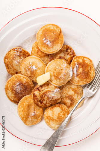 Dutch poffertjes mini pancakes are arranged on a white plate with red trim, dusted with sugar and topped with butter, while a fork rests along the right side in an overhead view