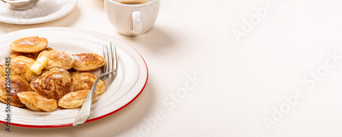 Small golden Dutch poffertjes pancakes topped with butter sit on a white plate with a red rim, while a fork and coffee cup rest nearby. Banner with copy space