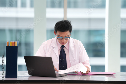 Asian man doctor reviewing document with laptop at bright medical office desk, focused professional expression during clinical research and healthcare administration