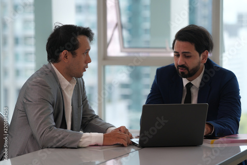 Business meeting with two men discussing work beside laptop in bright office, showing focused collaboration and professional communication during important workplace conversation