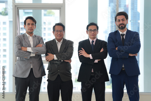 Business team portrait featuring four professional men in formal suits standing confidently in bright modern office with city buildings visible through large windows