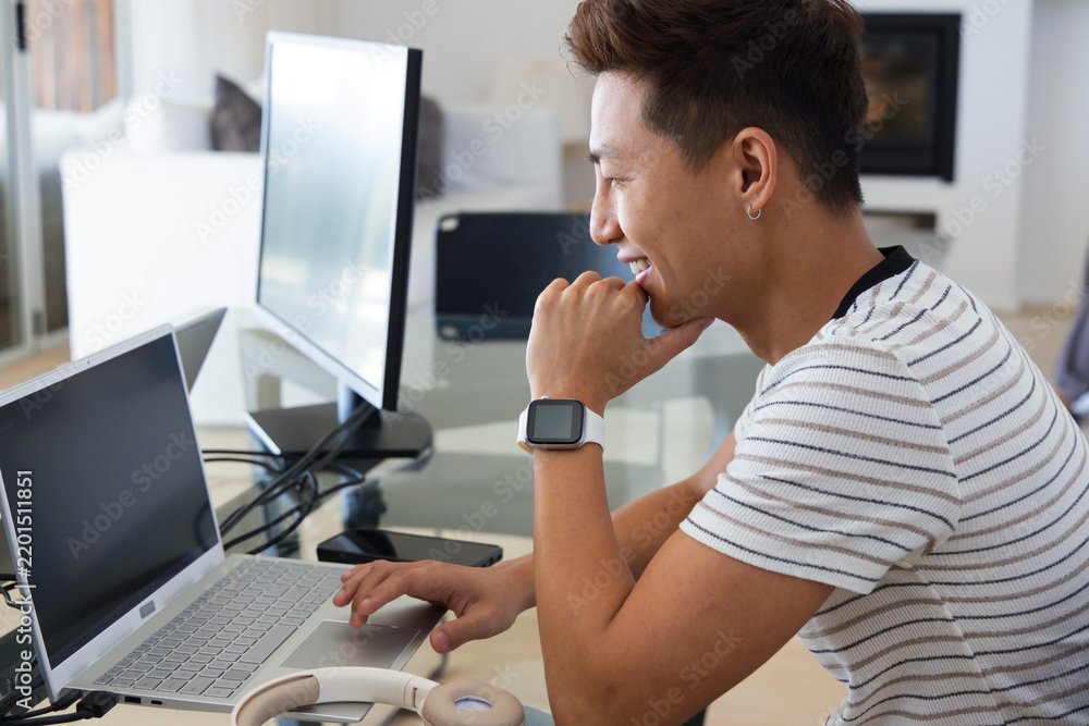 Fototapeta premium Asian man sitting at glass-topped desk working on laptop and monitor, smiling, copy space