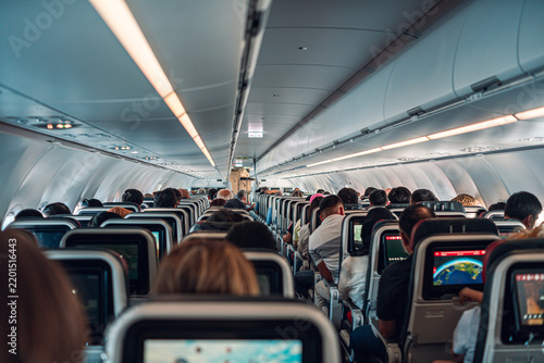 A packed airplane cabin shows passengers seated closely together, each engaged with their personal screens. The image captures the confined yet organized environment of air travel.