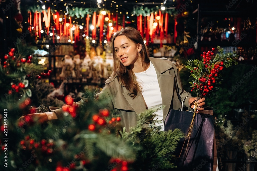 Obraz premium Woman choosing festive evergreen branches with red berries while shopping at a warmly lit seasonal market, wearing a coat and carrying bags among decorated holiday greenery