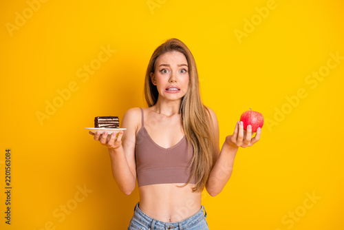 Obraz na plátně Young woman holding a cake slice and an apple, feeling a dilemma in choosing foo