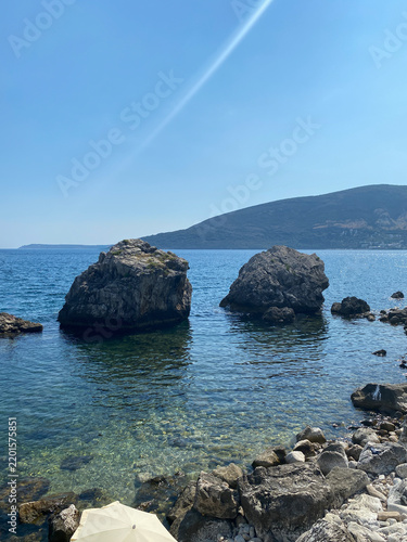 Two large rock formations stand in the clear, calm waters of Herceg Novi, Montenegro, near a rocky shoreline under a bright blue sky