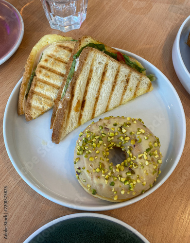 Grilled panini sandwich cut in half and a glazed pistachio donut are served on a white plate