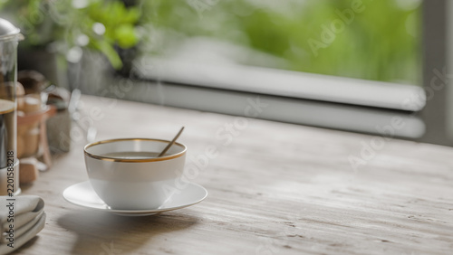 Steaming cup of coffee with gold rim on saucer beside French press on wooden table, blurred outdoor greenery visible through window in background.