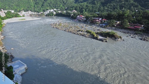 Currents move around a rocky gravel bar in the Ganga River, with wooded banks and buildings beyond in Rishikesh.