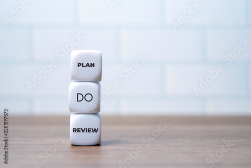 Stacked white dice featuring the words PLAN, DO, and REVIEW on a wooden desk. Business management cycles, strategic execution, workflow concept.