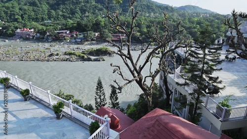 An elevated view of the Ganga River in Rishikesh includes white railings, conifers and a red roof in the foreground.