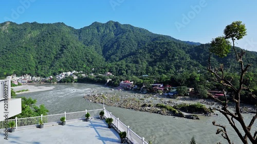 The Ganga River curves between green hills in Rishikesh, viewed across a white terrace railing and potted shrubs.