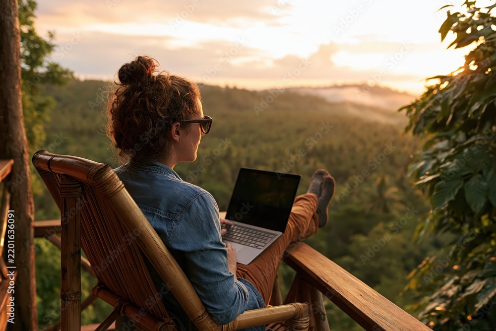 Obraz premium Woman using laptop on bamboo chair overlooking tropical forest at sunset