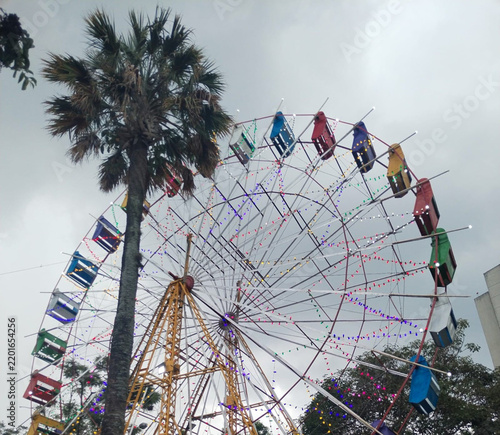 Fotografie Rueda de la fortuna iluminada con luces de colores junto a una palmera bajo un cielo nublado
