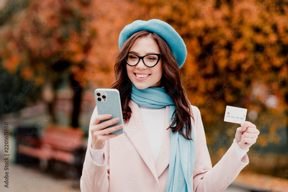 Obraz premium Young woman smiling while shopping online with smartphone and credit card in autumn park