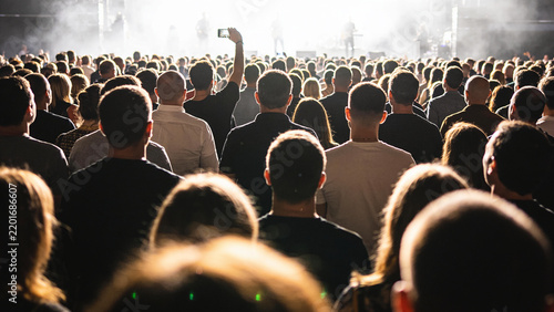 A large crowd of people at a concert, illuminated by stage lights, with one person in the foreground holding up a phone to record.