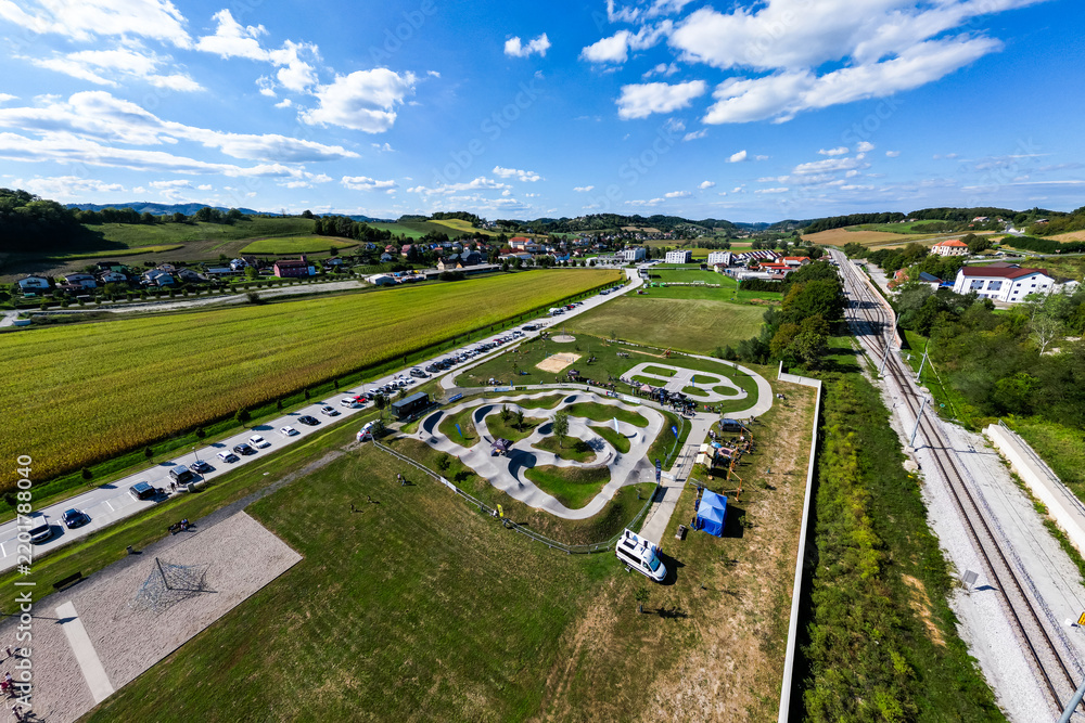 Obraz premium A wide aerial view of the asphalt pumptrack park in Pesnica pri Mariboru, Slovenia, surrounded by green fields, rolling hills, a railway line, and a bright blue sky filled with fluffy white clouds.