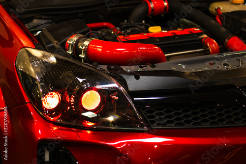 Red sports car front with illuminated headlights, black grille, glossy bodywork, and visible performance engine components in close up automotive workshop display
