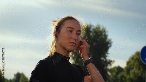 Young woman adjusting sunglasses and earbud outdoors in sunlit park with blurred trees and sky scene
