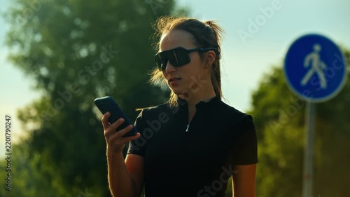Smiling woman with smartphone outdoors in sunlit park beside pedestrian sign and green trees backlit