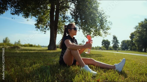 Young woman lifting water bottle while resting in sunny park beside large tree after outdoor workout