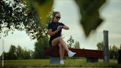 Athletic woman seated on park bench checks wristwatch beside trees under soft daylight in calm green