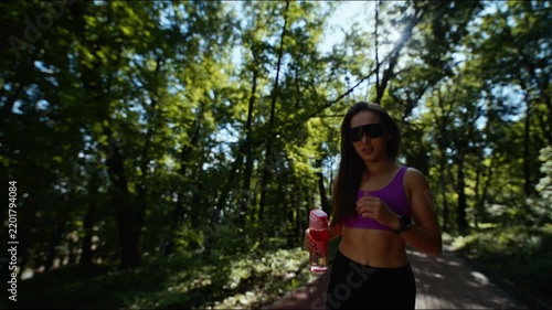 Young woman, resting with water bottle on sunlit forest path wearing sunglasses, and sportswear gear