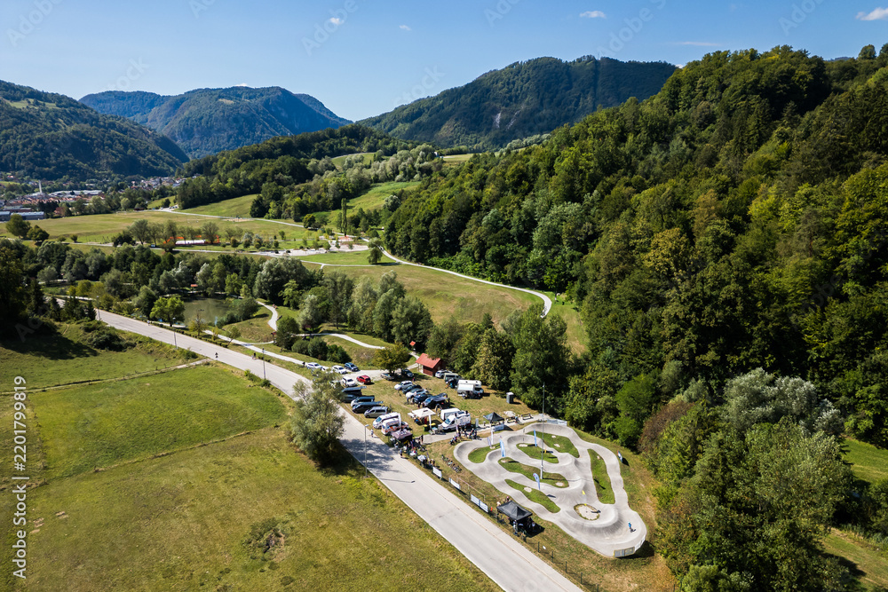 Fototapeta premium A wide aerial view of the asphalt pumptrack park in Zagorje ob Savi, Slovenia, surrounded by green fields, rolling hills, a railway line, and a bright blue sky filled with fluffy white clouds.