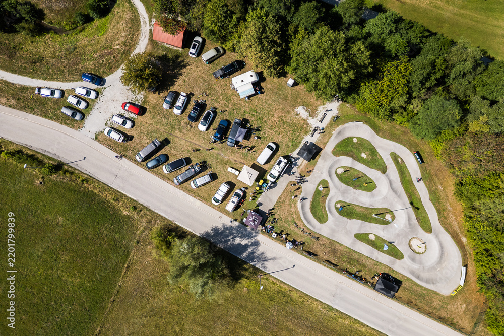 Obraz premium A wide aerial view of the asphalt pumptrack park in Zagorje ob Savi, Slovenia, surrounded by green fields, rolling hills, a railway line, and a bright blue sky filled with fluffy white clouds.