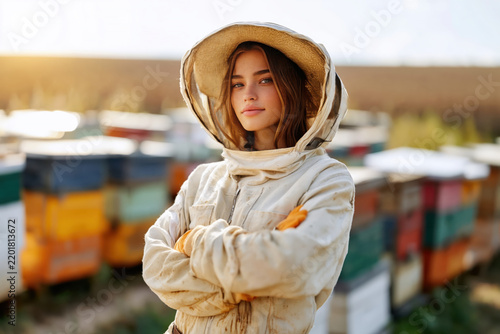 Confident young female beekeeper wearing protective suit standing by colorful beehives outdoors during sunset