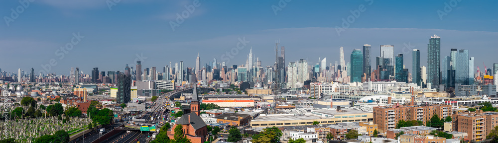 Obraz premium Aerial wide panorama captures the distant Manhattan skyscrapers rising beyond Queens neighborhoods, cemetery grounds, and highway traffic on a clear day.