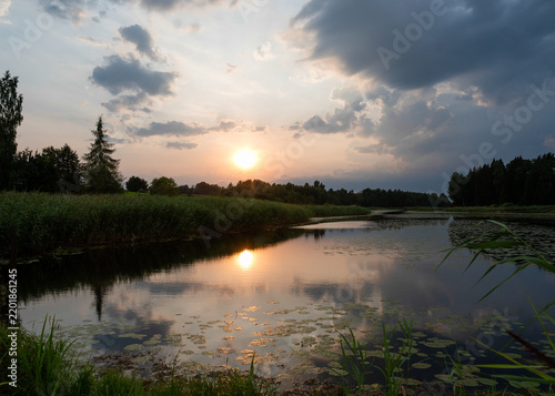 A magical landscape featuring a beautiful sunset or sunrise over a pond or lake on a summer evening; a cloudy sky in the background, with reeds and water reflections in the foreground;