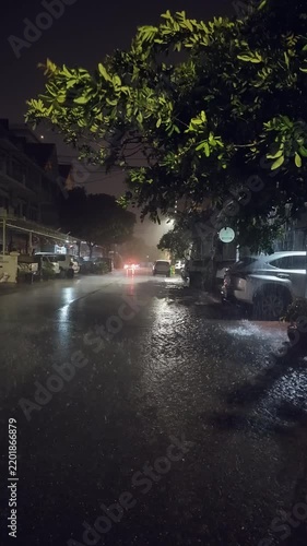 Vertical shot of a flooded small city street at night during a heavy rainstorm, with cars parked under lush trees while rain pours down.