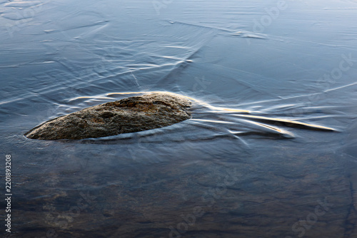 Bild auf Leinwand Close-up photograph of a stone emerging from dark frozen lake water with visible stress lines in the thin ice crust