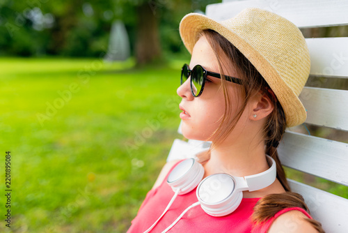 Profile portrait of a teenage girl wearing a straw hat, sunglasses, and white headphones around her neck, relaxing on a white wooden chair in a green park.