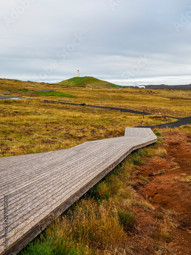 Wild Icelandic landscape with lava fields, local vegetation, wooden track, black pebble trail near Gunnuhver Hot Springs. In the distance visible Reykjanesviti Lighthouse. Reykjanes Peninsula, Iceland