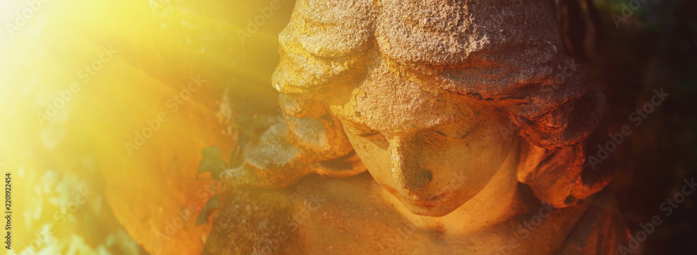 Fototapeta premium Ancient stone angel statue illuminated by radiant golden sunlight, symbolic image of heaven, divine light, faith, hope and eternal peace.