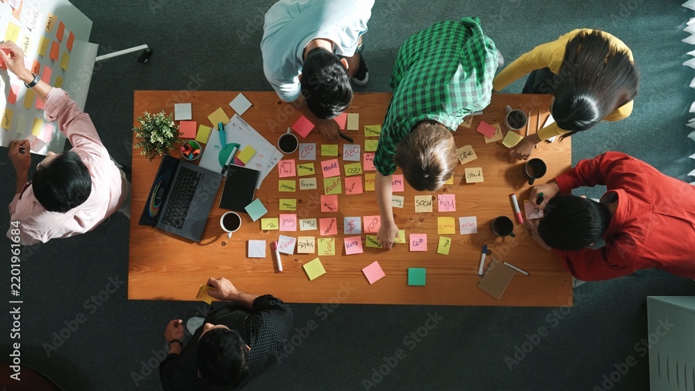 Obraz premium Top view of skilled business people discuss plan at meeting room while writing idea at sticky notes and stick at whiteboard. Group of diverse team working together to planning strategy. Convocation.