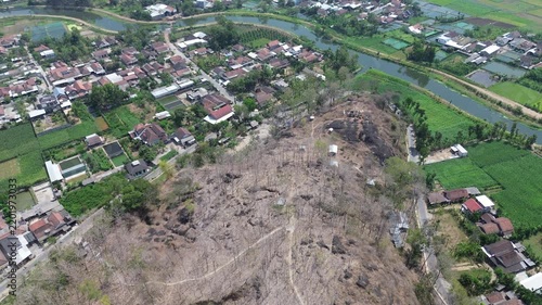 High angle drone shot of a dry rocky hill with sparse trees surrounded by a rural village, river canal, and green fields during dry season.