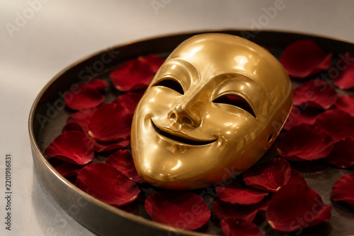 A single smiling gold mask resting on a tray of scattered rose petals.