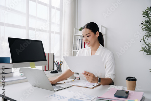 Focused Professional at Work: A diligent woman, illuminated by natural light, actively engages with documents and her laptop at a clean, organized desk, embodying concentration and productivity.