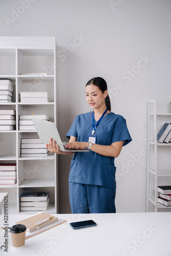 Nurse with Laptop: A focused female nurse in blue scrubs stands in a brightly lit office, engrossed in her work on a laptop, surrounded by bookshelves filled with medical literature.