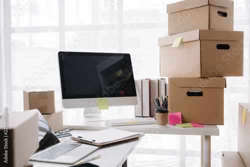 Office Relocation Chaos: A cluttered desk with boxes piled high, a computer monitor, and office supplies suggests a business in the midst of moving or organizing inventory.
