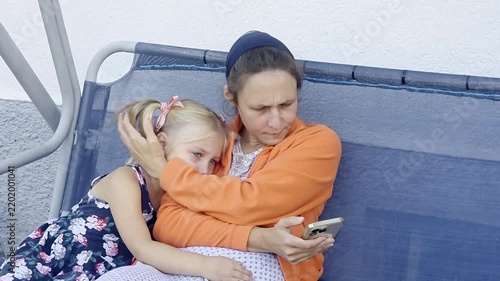 Mother hugging her little daughter while sitting together on a swing at a playground. The mother is using a smartphone while embracing her child. Family bonding, motherhood, childhood, parenting, mode