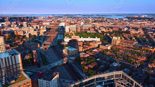 Highway and railway through Boston neighborhoods toward the harbor. Sunlit brick blocks flank parallel transport routes, with bridges, trees and islands beyond.