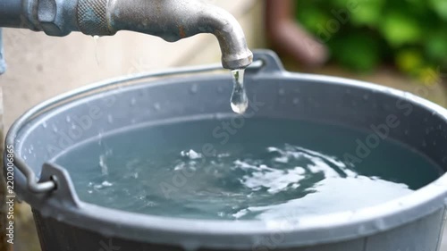 A solitary drop of fresh water drips from a metal faucet into a collection bucket, highlighting the critical need for global water conservation and sustainable resource management