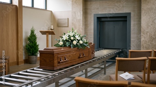 Wooden casket adorned with white floral arrangements on a mechanical roller track, preparing to enter a dark cremation chamber inside a committal or crematorium room