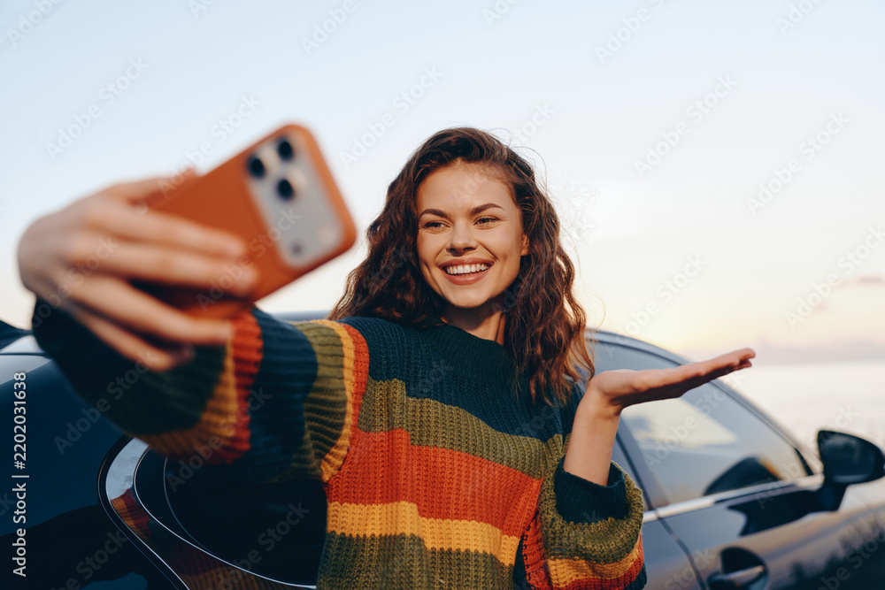 Obraz premium Smiling woman taking selfie with smartphone near car at sunset. Concept of modern lifestyle, travel, and mobile communication. Bright emotions and happy mood create a warm atmosphere outdoors.
