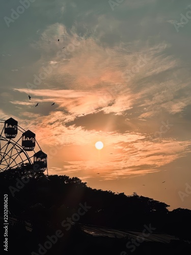 Stunning sunset view with ferris wheel silhouette against orange cloudy sky and flying birds flocks.