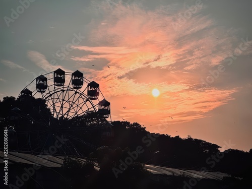 Silhouette of ferris wheel against dramatic sunset sky with golden clouds and flying birds in park.